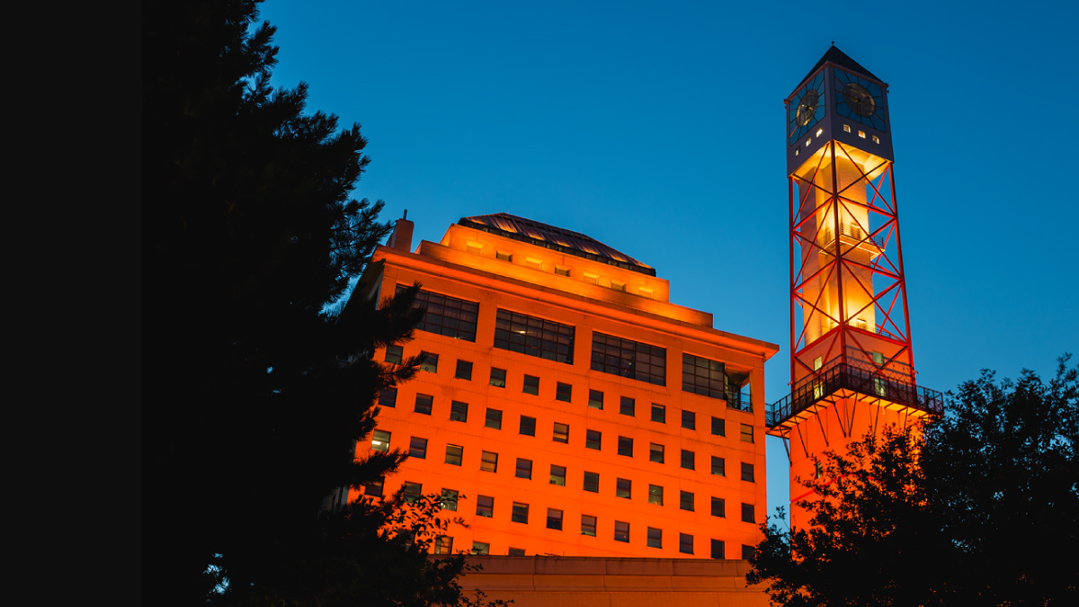 The Mississauga Civic Centre and clock tower light in the colour orange surrounded by trees in the dark night sky. 