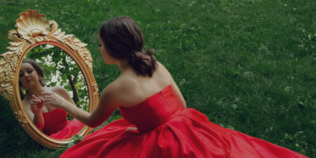 A woman in a red dress looking into a mirror.