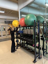 A woman at an indoor gym picking up a workout ball from the rack 