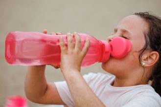 Girl with long dark hair wearing white T-shirt, drinking water from pink plastic bottle holding with hands. 