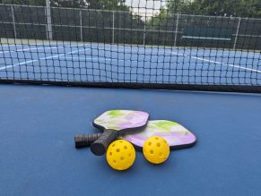 Pickleball equipment on the court including two racquets and two yellow outdoor pickleball balls