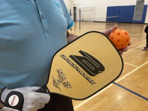 A person holding a pickleball racquet and ball getting ready to play in a gym