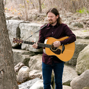 Matt Zaddy playing a guitar by a river.