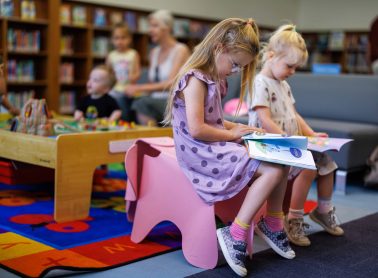 Two young girls sitting on small chairs in a library, reading picture books. Other children and an adult are visible in the background near a play area.