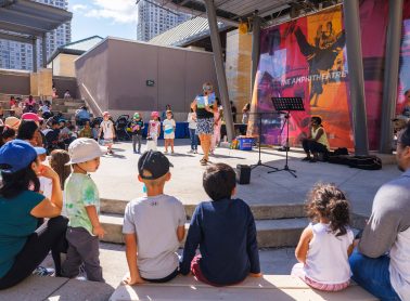 An outdoor storytime event with children and adults gathered around a storyteller reading from a book. The event is held in an amphitheater with a colorful backdrop and buildings in the background.