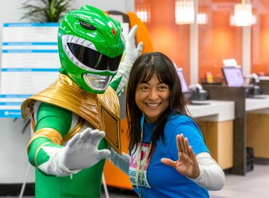 A woman smiling and posing with a person dressed in a green superhero costume, both striking a playful action pose. The background shows a brightly lit indoor setting with counters and computer terminals.