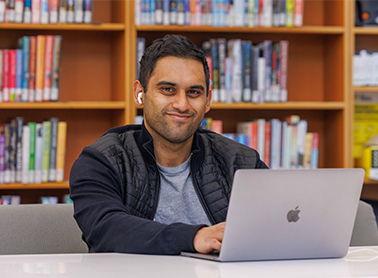 A man sitting at a table in a library, smiling and working on a laptop with bookshelves in the background.