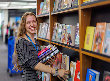 A woman with long hair, smiling and holding a stack of books, standing in front of bookshelves in a library.