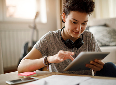 A young person with short hair, concentrating on a tablet, wearing headphones around their neck while sitting at a desk with study materials.