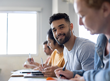 A group of students sitting at a table, focused on their work, with one man smiling at the camera while others write or study materials in front of them.