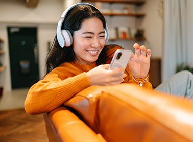 A woman wearing headphones, smiling, and looking at her phone while lounging on a couch.