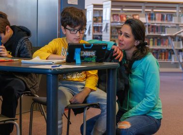 A woman in a blue shirt kneels down beside a child looking at a digital screen.