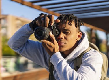 A young man in a grey hoodie holds a telescoping camera up to his eye.