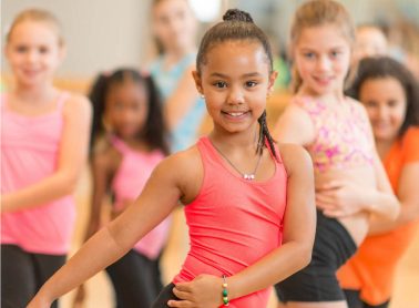 A group of smiling girls in colourful outfits pose confidently in a dance class, with one girl in a bright pink tank top at the front.